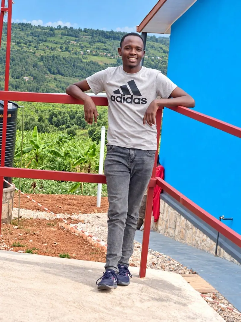 Alphonse, Lea's son, a young man in a grey t-shirt leaning on a red railing, green hills behind.