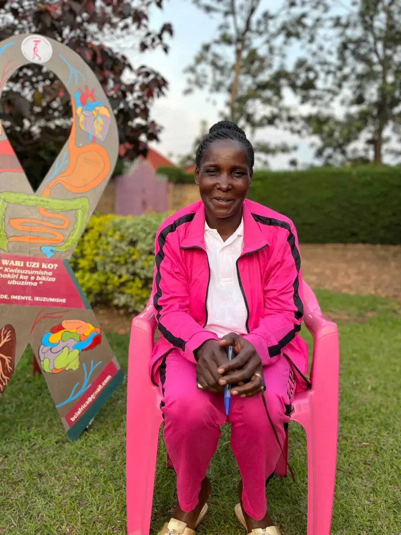 Speciose, a woman in a pink tracksuit, seated on a pink chair outdoors beside a painted breast-health ribbon board.